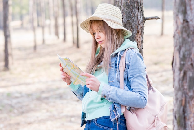 Feminino viajante com sua mochila em pé debaixo da árvore, olhando para o mapa
