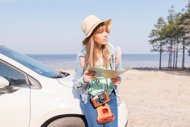 Feminino viajante inclinando-se perto do carro branco segurando o mapa na mão, olhando para longe