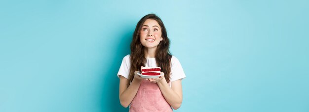 Feriados e celebração aniversariante sonhadora fazendo desejo e olhando esperançoso segurando bolo de aniversário e sorrindo em pé sobre fundo azul