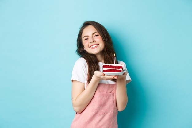 Feriados e comemorações. Linda garota glamour comemorando o aniversário dela, segurando o prato com bolo e sorrindo alegre, comemorando, em pé sobre um fundo azul.