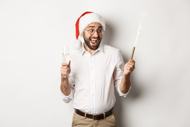 Férias de inverno e celebração. Homem animado celebrando a véspera de Ano Novo com brilhos de fogos de artifício e bebendo champanhe, usando chapéu de Papai Noel