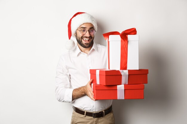 Férias de inverno e celebração. Um cara feliz trazendo presentes de natal, segurando presentes e usando chapéu de Papai Noel, em pé