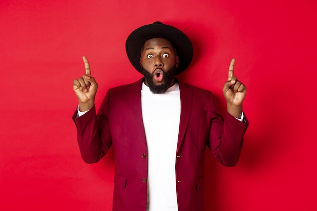 Férias de inverno e conceito de compras. homem afro-americano impressionado com roupa de festa apontando os dedos para cima, conferindo a promoção de natal, em pé sobre um fundo vermelho