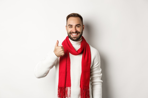 Férias de inverno e conceito de compras. homem bonito confiante aparecendo o polegar, de pé na camisola de natal e lenço vermelho, fundo branco.