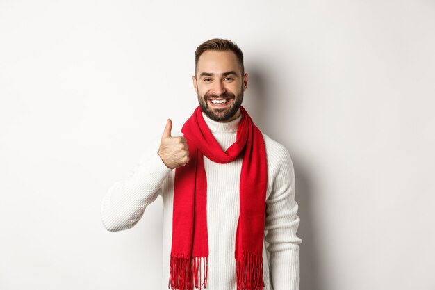 Férias de inverno e conceito de compras. Homem bonito confiante aparecendo o polegar, de pé na camisola de Natal e lenço vermelho, fundo branco.