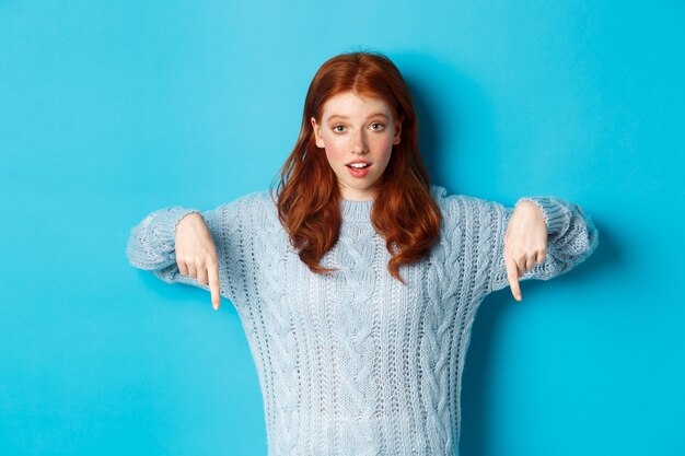 Férias de inverno e conceito de pessoas. Linda garota adolescente apontando o dedo para baixo, mostrando um anúncio, em pé sobre um fundo azul