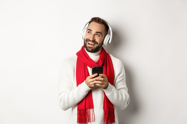 Férias de inverno e conceito de tecnologia. Homem feliz ouvindo podcast de música em fones de ouvido, segurando o telefone celular e olhando para o espaço vazio, fundo branco