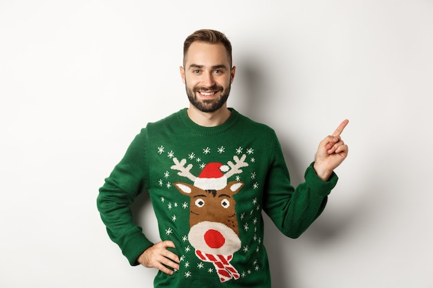 Férias de inverno e natal. Homem adulto barbudo e bonito apontando o dedo para o banner promocional, usando um suéter engraçado contra um fundo branco