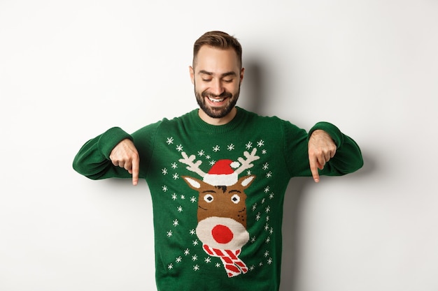 Férias de inverno e natal. Homem caucasiano feliz com suéter verde, apontando o dedo para baixo e parecendo satisfeito, em pé sobre um fundo branco