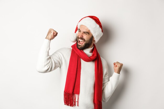 Férias de inverno e o conceito de festa de ano novo. Bonito homem barbudo com chapéu de Papai Noel ganhando o prêmio, atingir o objetivo e celebrar, fazendo o punho bombar e dizendo que sim, fundo branco.