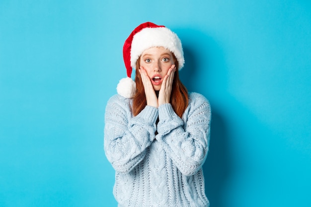 Férias de inverno e o conceito de véspera de Natal. Ruiva surpresa com chapéu de Papai Noel, olhando para a câmera com descrença, boca aberta e maravilhada, em pé sobre um fundo azul