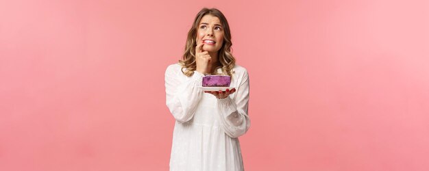 Férias de primavera e conceito de festa Retrato de uma jovem loira hesitante e alarmada pensando hesitando em comer ou não uma sobremesa deliciosa com calorias em dieta resistir à tentação