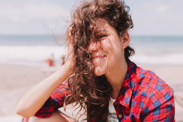 Férias de verão. Mulher feliz e agradável com cabelos ondulados voando, sentada na costa do oceano