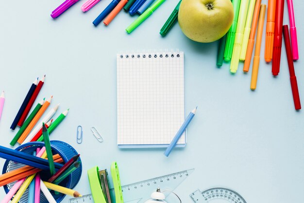 Ferramentas de desenho coloridas espalhadas em torno do bloco de notas em branco na mesa azul