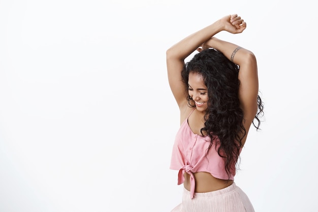 Festa divertida e conceito de beleza mulher afro-americana atraente de cabelos encaracolados em top rosa com tatuagens dançando alegremente fechando os olhos e levantando as mãos despreocupadamente apreciando o fundo branco da festa