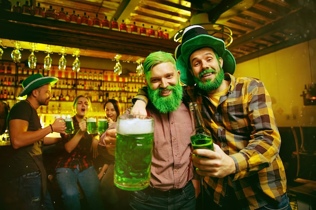 Festa do dia de são patrício. amigos felizes está comemorando e bebendo cerveja verde. homens e mulheres jovens usando chapéus verdes. pub interior.