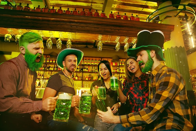 Festa do dia de são patrício. amigos felizes estão comemorando e bebendo cerveja verde. homens e mulheres jovens usando chapéus verdes. pub interior.