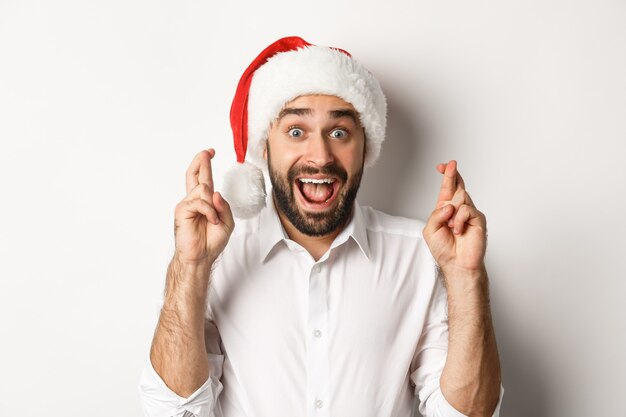 Festa, férias de inverno e conceito de celebração. Homem feliz com chapéu de Papai Noel fazendo um desejo de Natal, cruze os dedos para dar sorte e parece animado