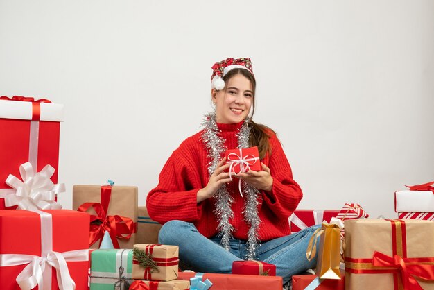 Festeira de vista frontal com chapéu de Papai Noel segurando um presente com as duas mãos sentadas em volta dos presentes