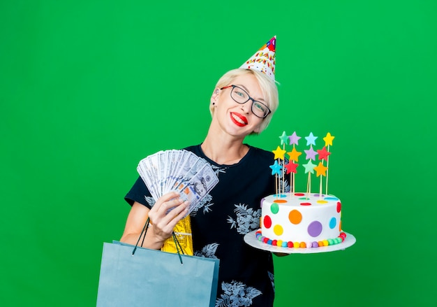 Foto grátis festeira jovem sorridente de óculos e boné de aniversário segurando um bolo de aniversário com estrelas, caixa de presente de dinheiro e um saco de papel, olhando para a câmera, isolada em um fundo verde com espaço de cópia