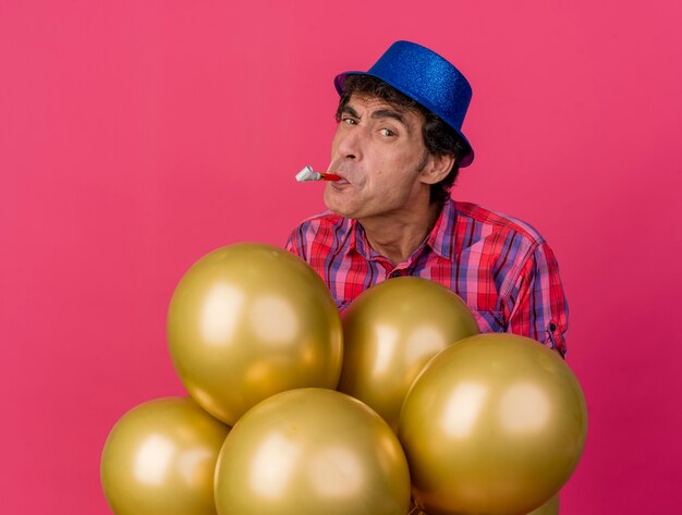 Festeiro duvidoso de meia-idade usando chapéu de festa em pé atrás de balões olhando para frente com um soprador de festa na boca isolado na parede carmesim
