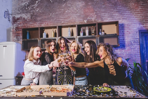 Festive mulheres amigas tilintando óculos na cozinha