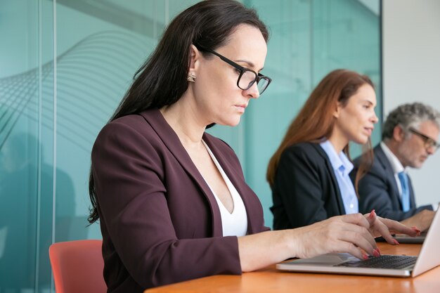 Fila de executivos sérios usando laptops, sentados em uma mesa e digitando