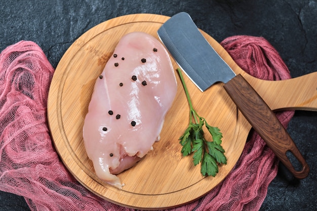 Filé de frango cru na placa de madeira com verduras e faca.