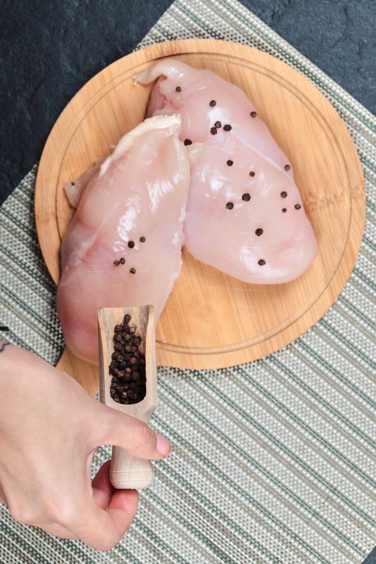 Foto grátis filé de frango cru na placa de madeira com verduras e mão segurando a colher de grãos de pimenta.