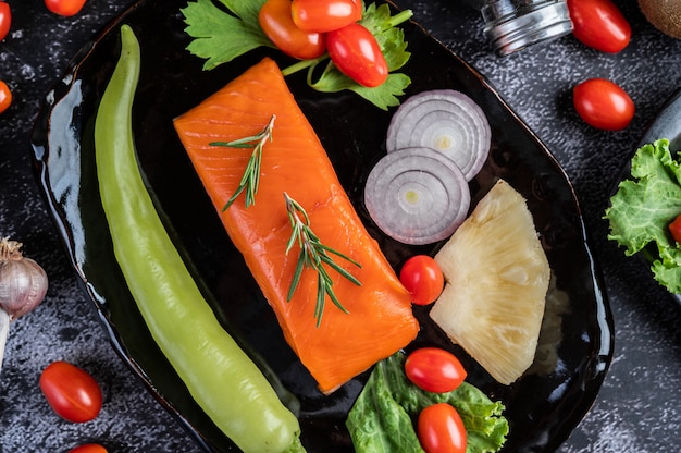 Filetes de salmão cru, pimenta, kiwi, abacaxi e alecrim em um prato e piso de cimento preto.