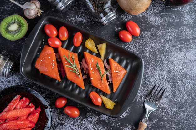 Filetes de salmão cru, pimenta, kiwi, abacaxi e alecrim em um prato e piso de cimento preto.