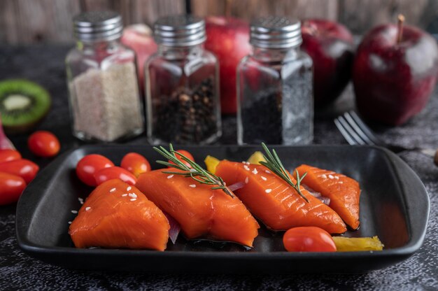 Filetes de salmão cru, pimenta, kiwi, abacaxi e alecrim em um prato e piso de cimento preto.