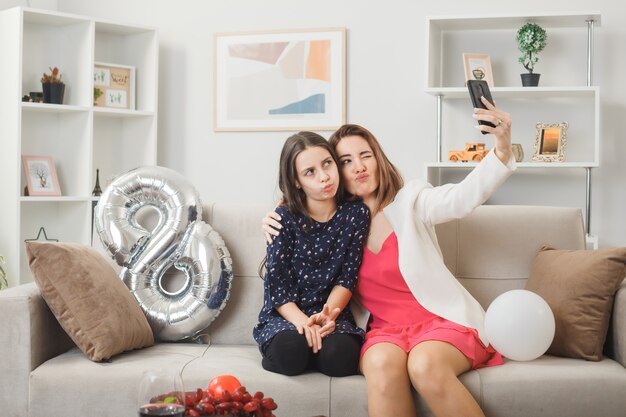 Filha e mãe satisfeitas no feliz dia da mulher, sentadas no sofá, tirando uma selfie na sala de estar