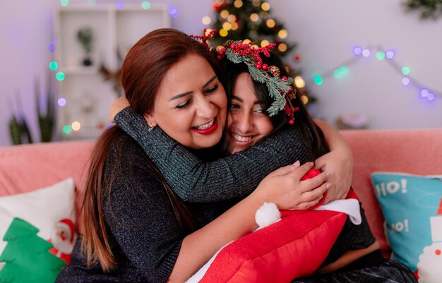 Filha e mãe sorridentes se abraçam sentadas no sofá, aproveitando o Natal em casa