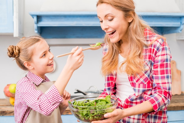 Filha sorridente, alimentando a salada para a mãe na cozinha