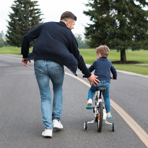 Filho andando de bicicleta no parque ao lado do pai por trás