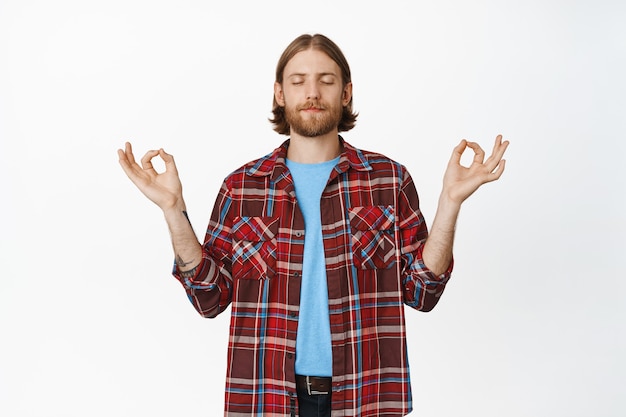 Fique calmo. homem loiro barbudo calmo e tranquilo fecha os olhos, meditando, segurando as mãos de lado no om, gesto do zen nirvana, praticando ioga no branco