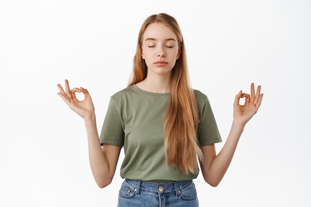 Fique calmo. Jovem relaxada meditando, fecha os olhos e dá as mãos de lado em sinal de mudra zen, pratica ioga, respirando e descansando a mente, em pé sobre um fundo branco