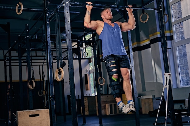 Fisiculturista loira com perna ferida em um curativo fazendo pull ups em cross fit stand em uma academia.