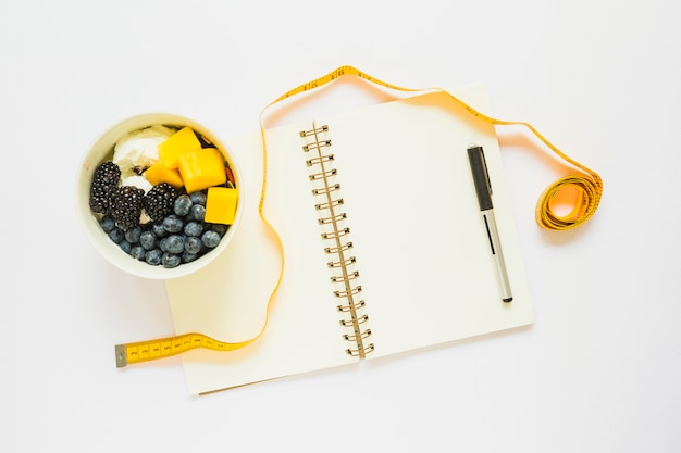 Foto grátis fita métrica; copo de frutas com iogurte; caneta e caderno espiral em fundo branco