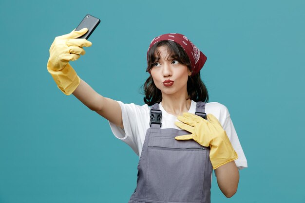 Flirty jovem faxineira vestindo bandana uniforme e luvas de borracha esticando o celular mantendo a mão no peito fazendo gesto de beijo tomando selfie isolada em fundo azul