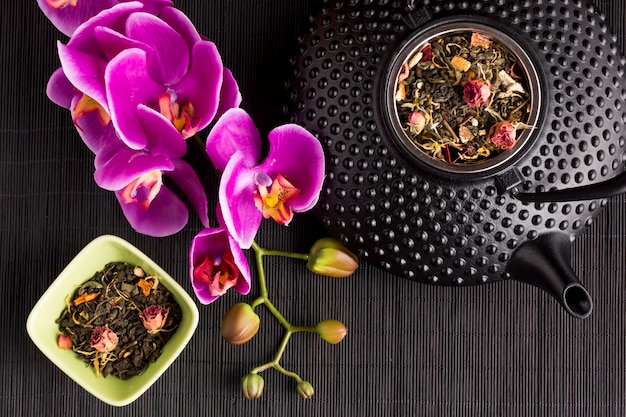 Flor de orquídea rosa e chá seco erva com textura bule de cerâmica em preto placemat
