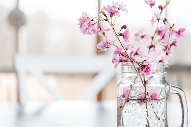 Flores de cerejeira e galhos em um copo d'água em cima da mesa sob as luzes