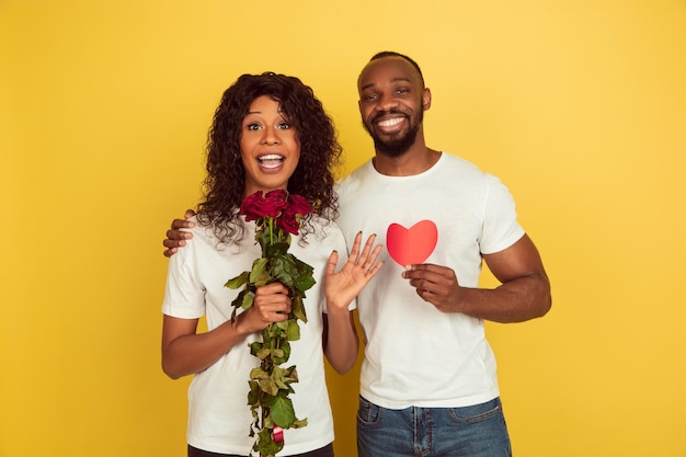 Flores e coração. Celebração do dia dos namorados, feliz casal afro-americano isolado no fundo amarelo do estúdio.