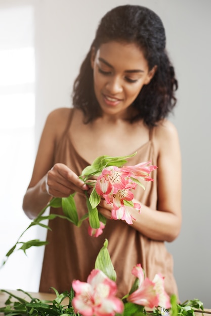 Florista africana bonita alegre que sorri fazendo o ramalhete no local de trabalho sobre a parede branca.