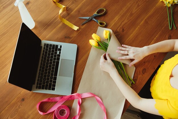 Florista no trabalho: mulher mostra como fazer buquê com tulipas. Jovem mulher caucasiana dá workshop online de fazer um presente, um presente para a celebração. Trabalhar em casa enquanto conceito isolado, em quarentena.