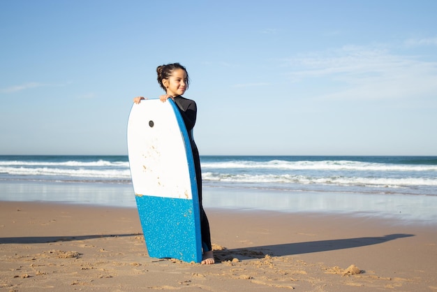 Foto grátis focada menina com prancha de surf na praia. criança morena em uniforme de surf em dia ensolarado, olhando de soslaio. esporte, lazer, conceito de estilo de vida ativo