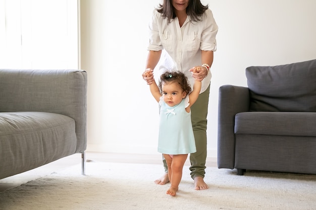 Focada menina no vestido azul claro, segurando as mãos da mãe e tentando andar em casa. Comprimento total. Conceito de paternidade e infância