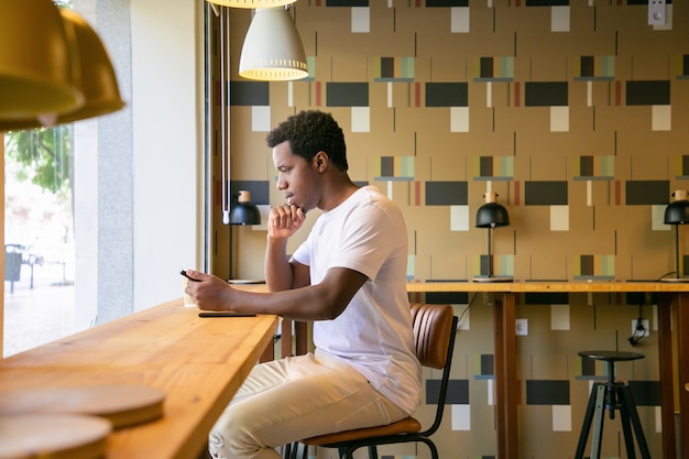 Focado jovem afro-americano sentado à mesa em um espaço de trabalho ou café, usando um tablet