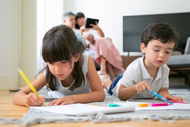 Focado menino e menina deitados no chão e desenhando na sala de estar enquanto os pais sentam juntos